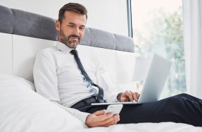 Businessman using phone and laptop while sitting on bed at home